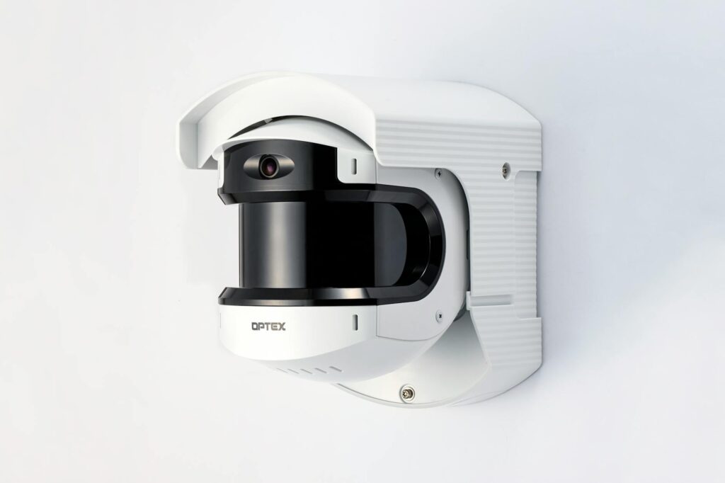 A wireless outdoor security camera mounted on the side of a house with a blurred suburban background